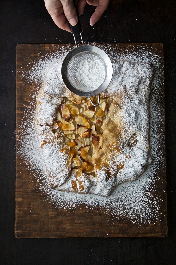 An apple tart on a chopping board being dusted with icing sugar