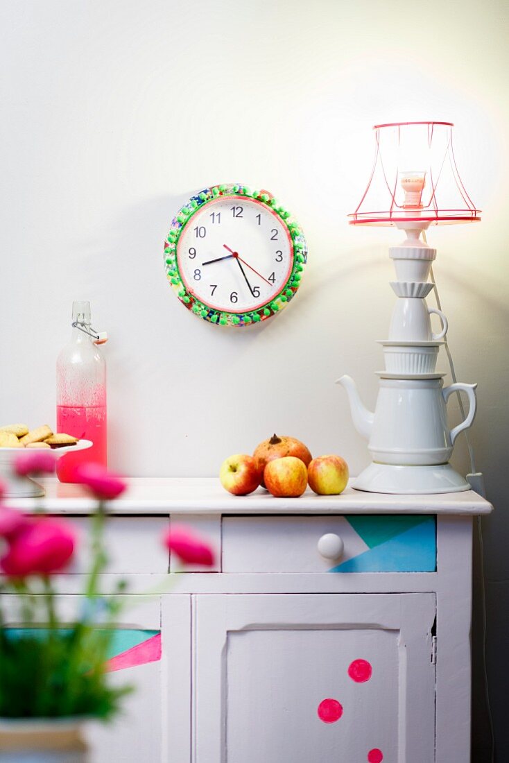 Wall clock decorated with colourful fabrics and neon green pompoms, vintage sideboard and table lamp made from stacked porcelain crockery