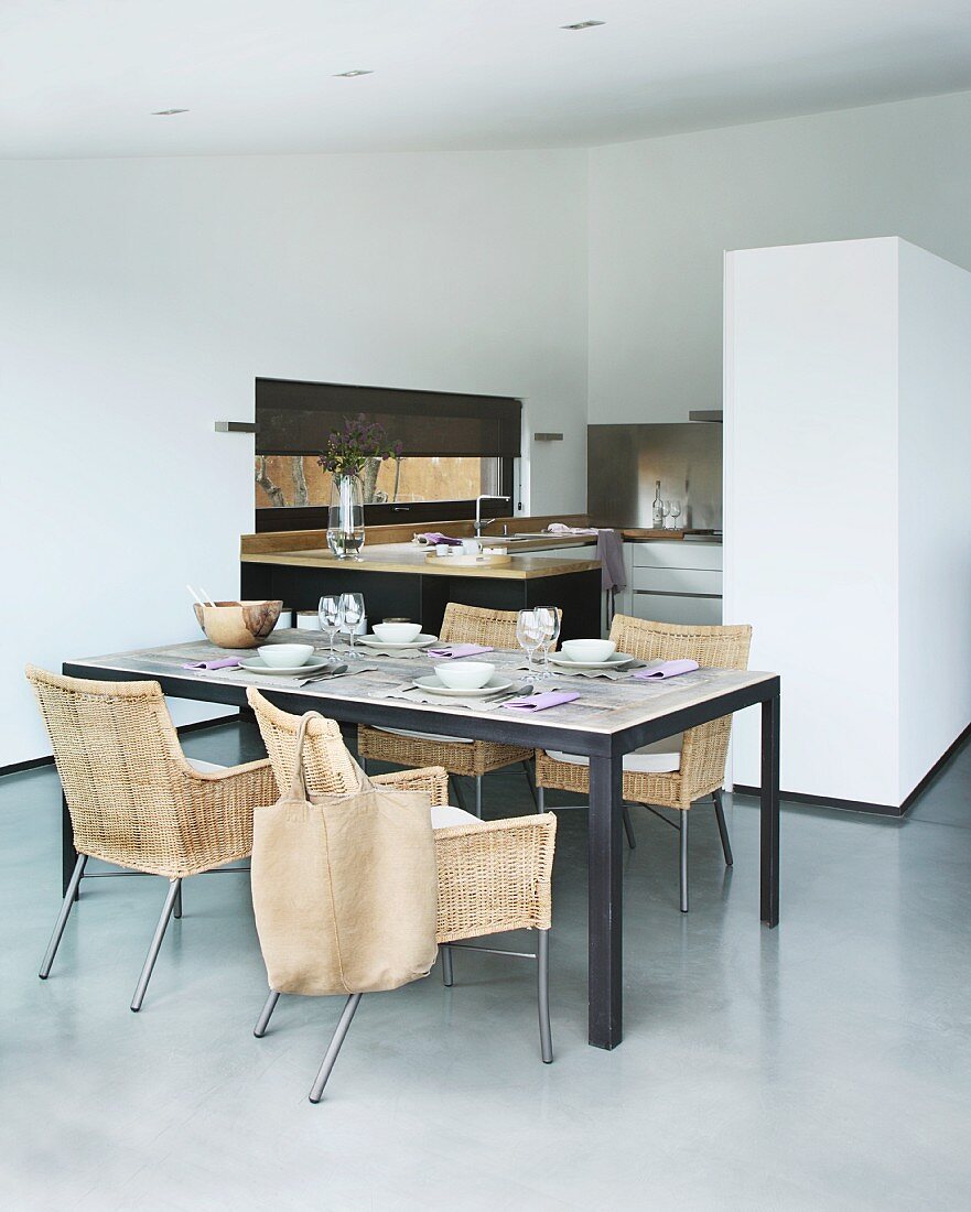 Set dining table and modern wicker chairs in purist interior; cabinet in partition wall and open-plan kitchen in background
