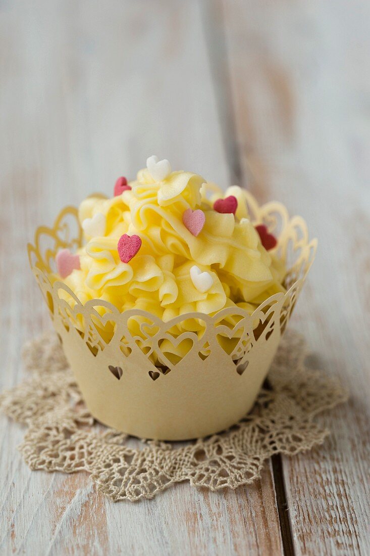 A cupcake decorated with yellow butter cream and sugar hearts on a crocheted doily