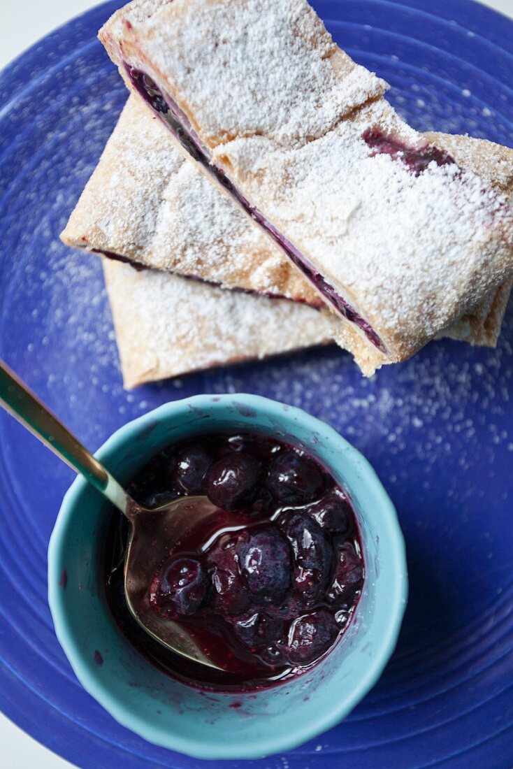 Three slices of blueberry strudel dusted with icing sugar with a bowl of blueberry sauce