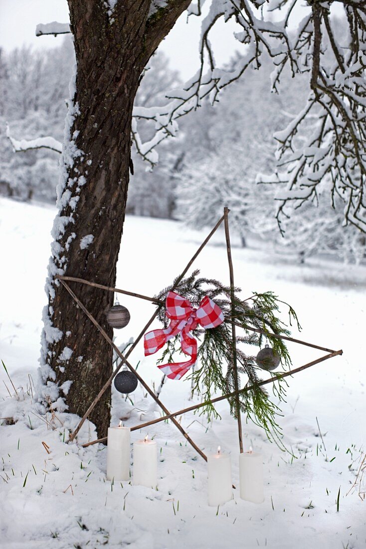 Star made from twigs decorated with baubles and ribbon leaning against tree and lit candles in snowy landscape