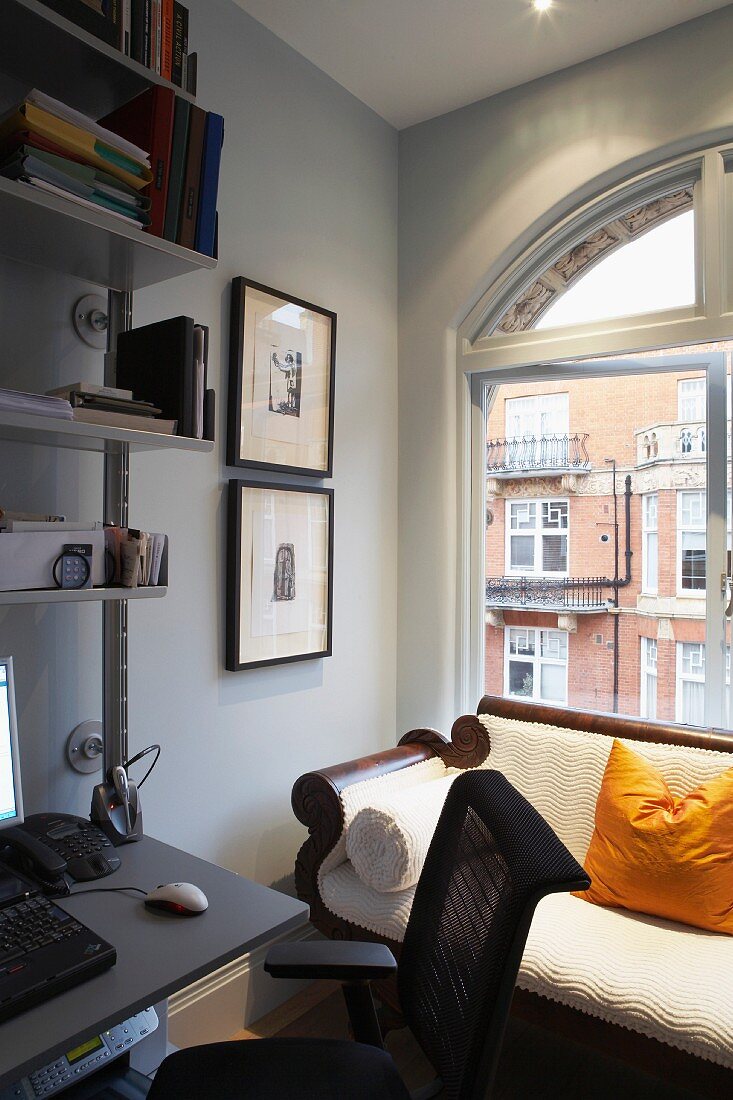 Antique couch with pale upholstery in front of arched window; modern desk integrated into metal shelving