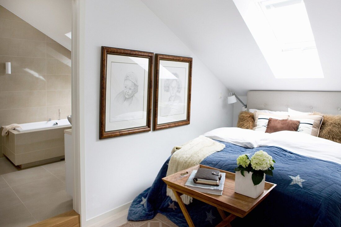 Doppelbett unter Dachfenster, seitlich offene Tür und Blick ins moderne Bad mit hellbeigen Fliesen
