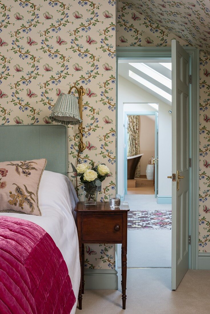 Romantisches Schlafzimmer mit Blümchentapete und antikem Nachttisch, Blick durch offene Tür