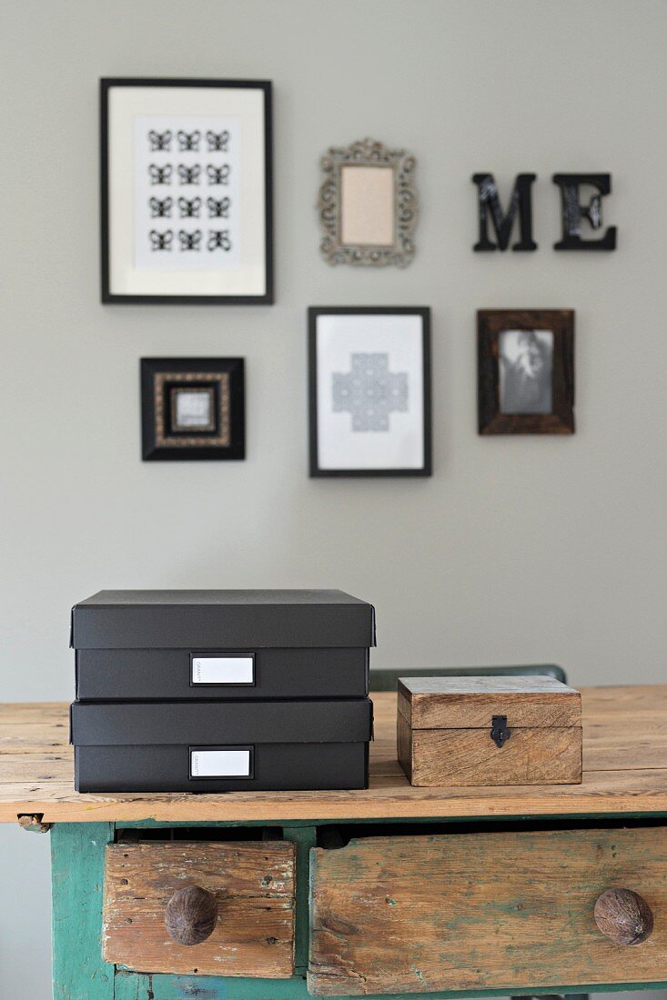 Schwarzer Ordnungsboxen mit Schild und Holzkiste auf Tisch, im Hintergrund gerahmte Bilder an grauer Wand