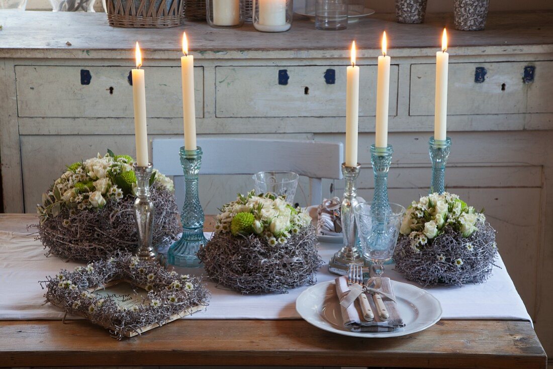 Romantic wreath of waxflowers, green chrysanthemums and cream polyantha roses amongst lit candles in candlestick on rustic table