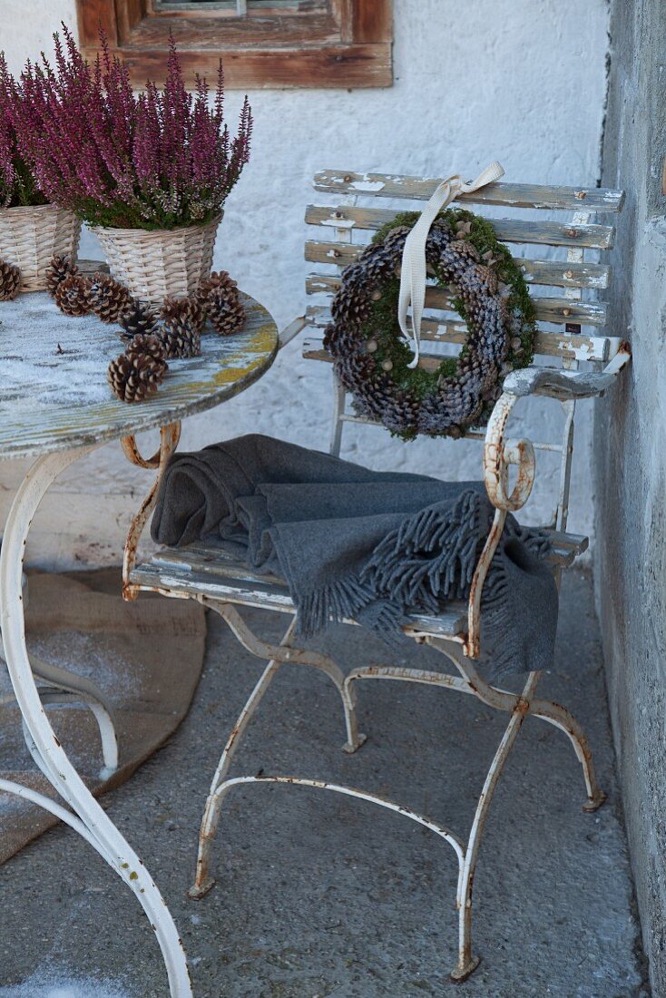 Grey blanket on vintage garden chair with peeling paint and wreath of pine cones on backrest