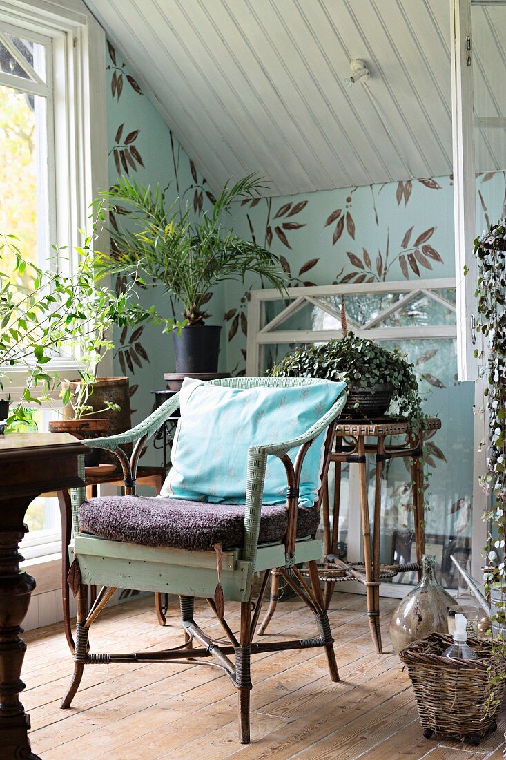 Armlehnstuhl aus Rattan in nostalgisch gestaltetem Dachzimmer, Pflanzentöpfe auf Blumenständer vor tapezierter Wand mit Blättermotiv