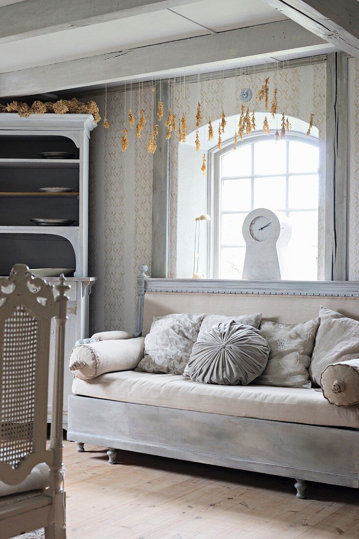 Old couch with upholstery and cushions in various shades of grey below window