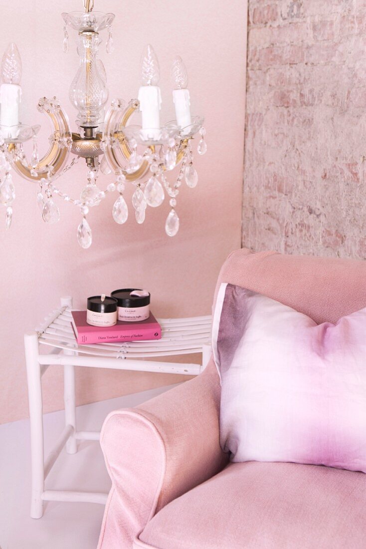 Pink armchair with scatter cushion and side table below crystal chandelier against pink-painted wall