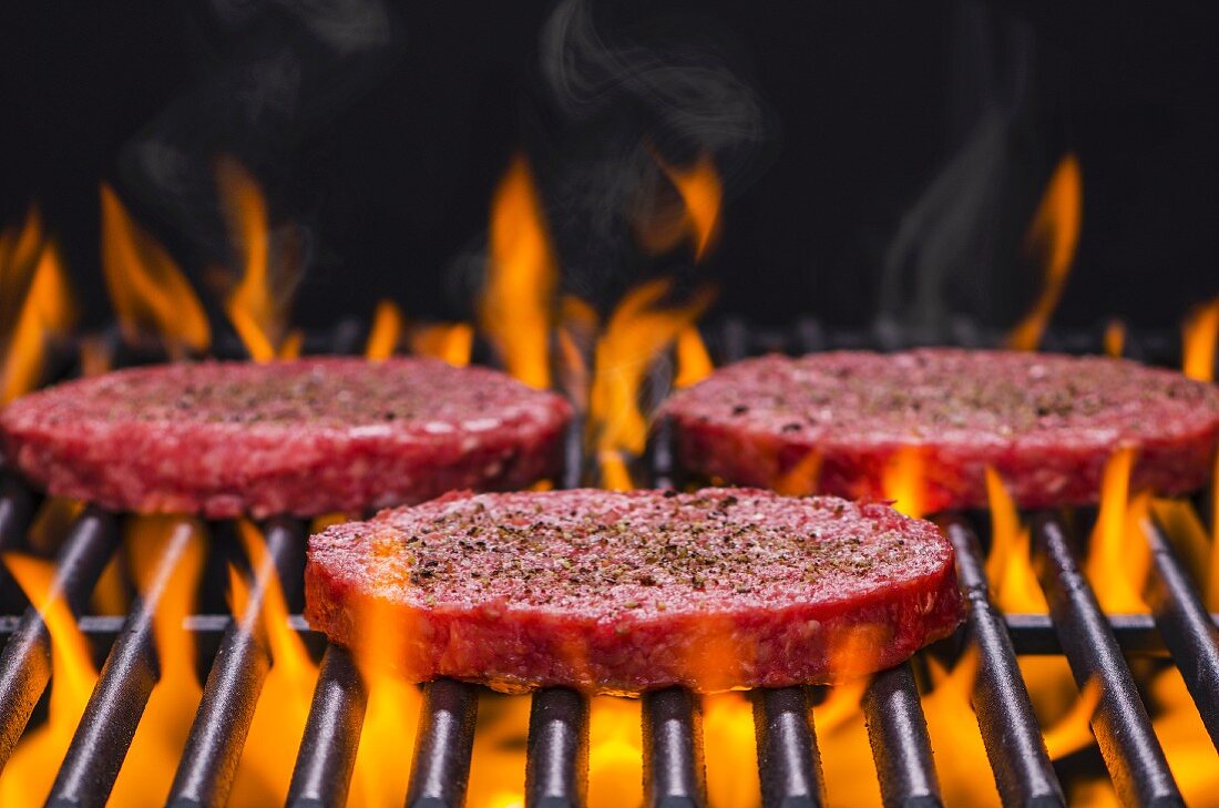 Raw hamburgers seasoned with salt and pepper on a flaming barbecue
