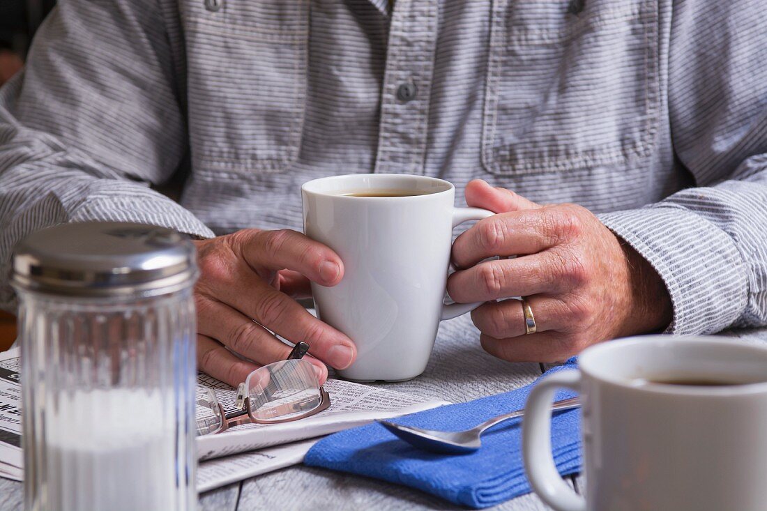 A man holding a cup of coffee and with a newspaper and a pair of glasses next to him