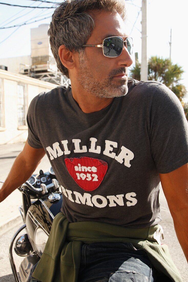 Mann in Jeans, T-Shirt und Sonnenbrille sitzt seitlich auf Motorrad