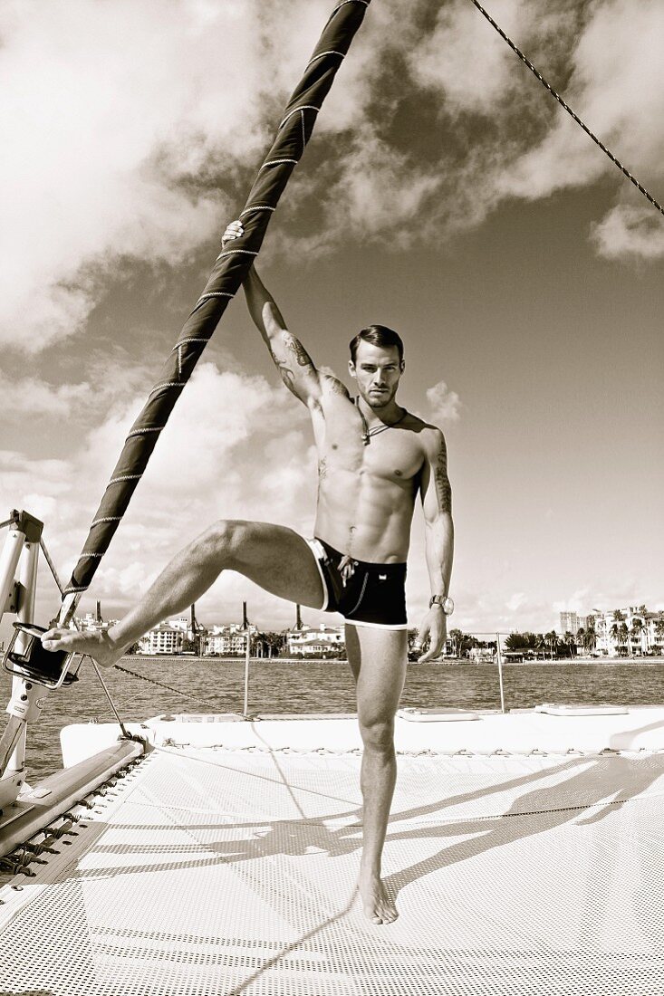 A young, athletic man with a side parting standing on a yacht wearing bathing shorts