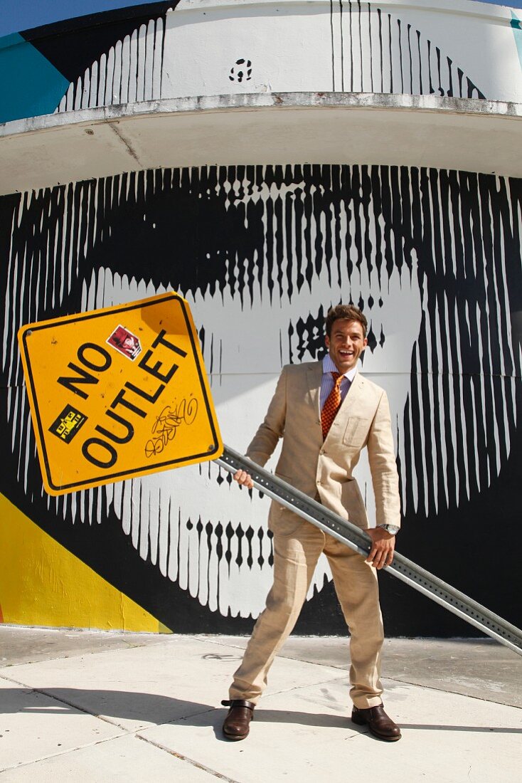 A young man wearing a light suit holding a 'NO OUTLET' street sign