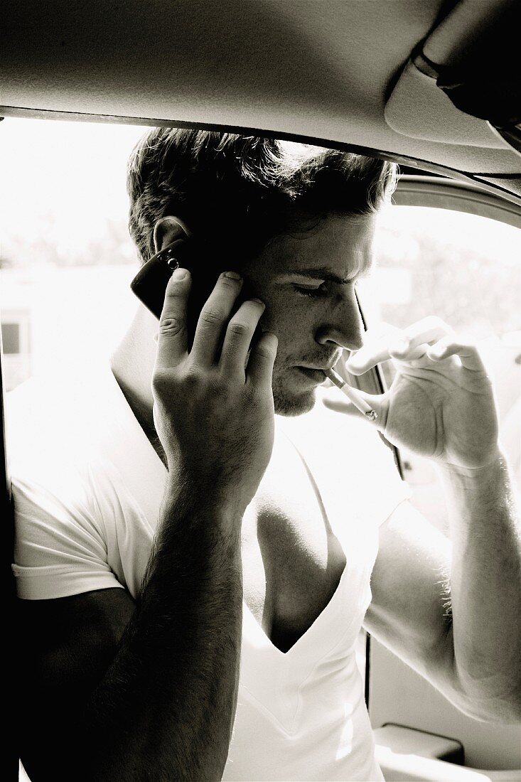 A young man with a cigarette in the mobile phone by a car