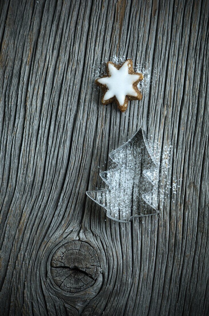 A Christmas tree-shaped cutter with icing sugar and cinnamon stars on a wooden board