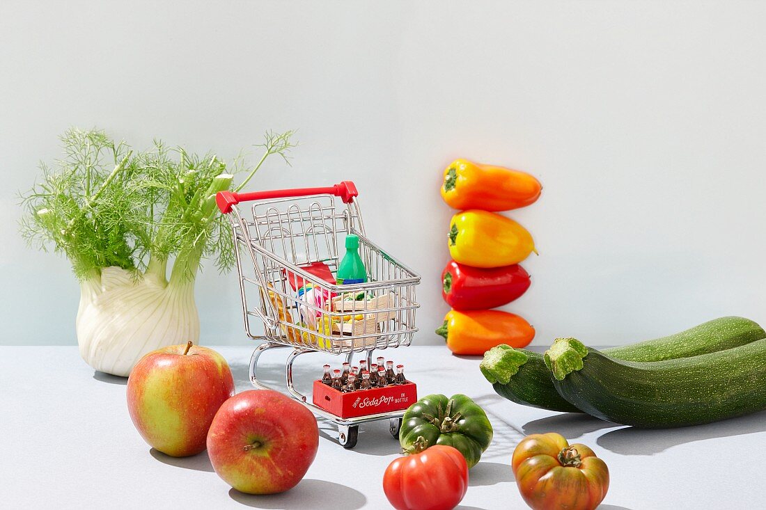 A mini shopping trolley filled with toy foodstuffs next to fresh fruit and vegetables