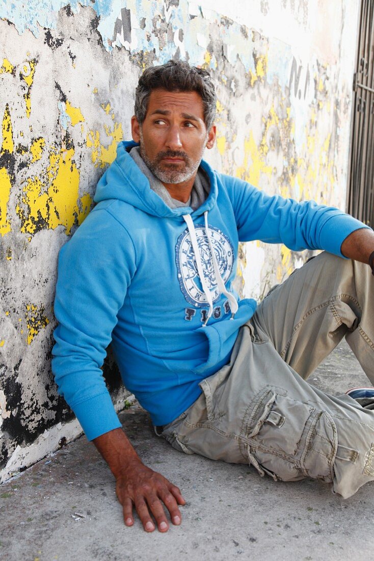 Mann mit hellblauem Kapuzensweater lehnt sich sitzend an Strassenmauer