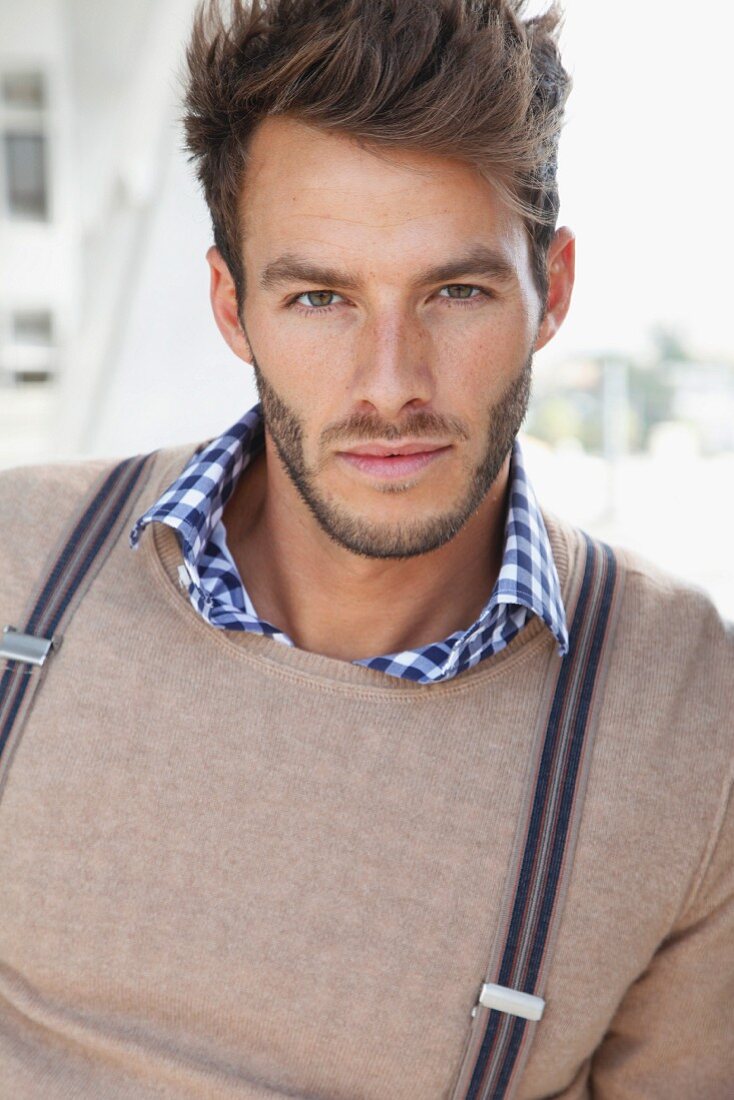 A young man wearing a checked shirt, a sand-coloured jumper and braces