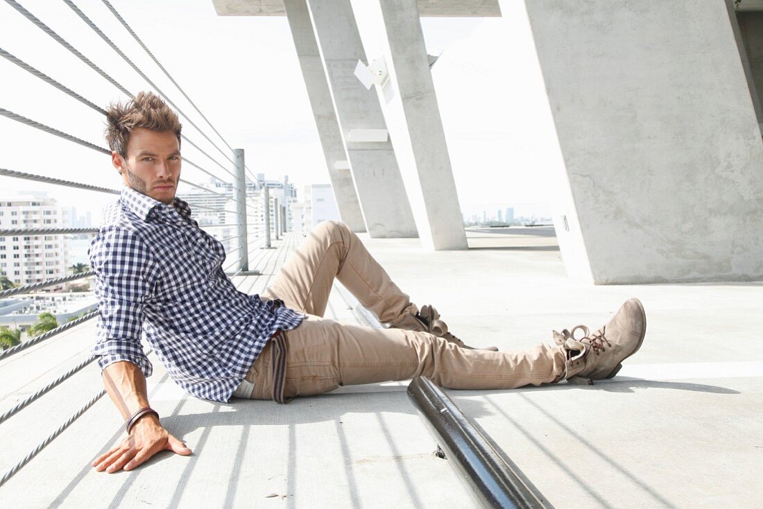 Junger Mann mit kariertem Hemd und sandfarbenem Jeans auf dem Boden sitztend
