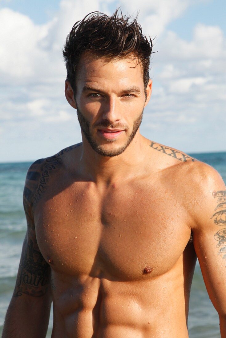 A young, topless, tattooed man by the sea