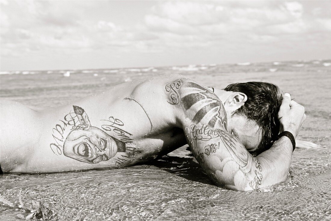 Junger, tätowierter Mann mit nacktem Körper am Meer im Sand liegend