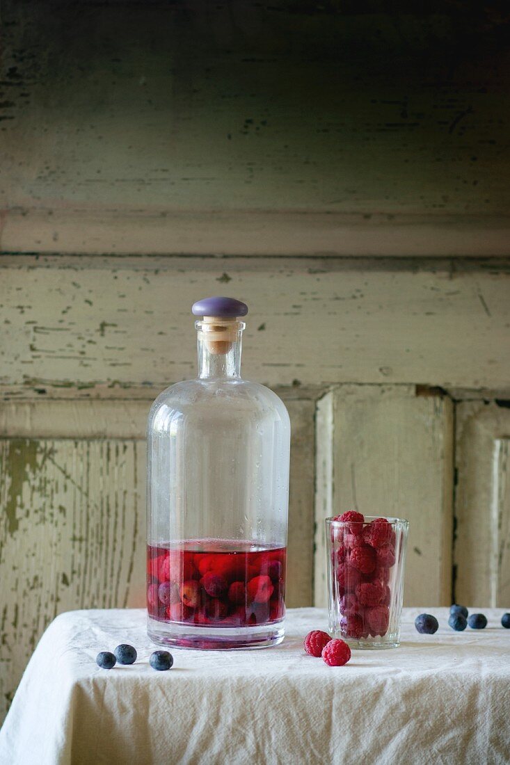 Eine Flasche hausgemachter Beerenlikör mit Erdbeeren und Heidelbeeren auf Tisch mit weißem Tischtuch
