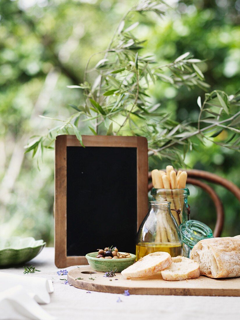 Ciabatta, Oliven, Olivenöl, Grissini und Schiefertafel auf Gartentisch