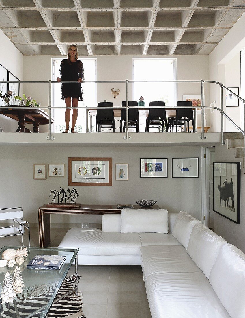 Open-plan interior with white corner sofa, woman on gallery with metal balustrade and dining area below concrete coffered ceiling