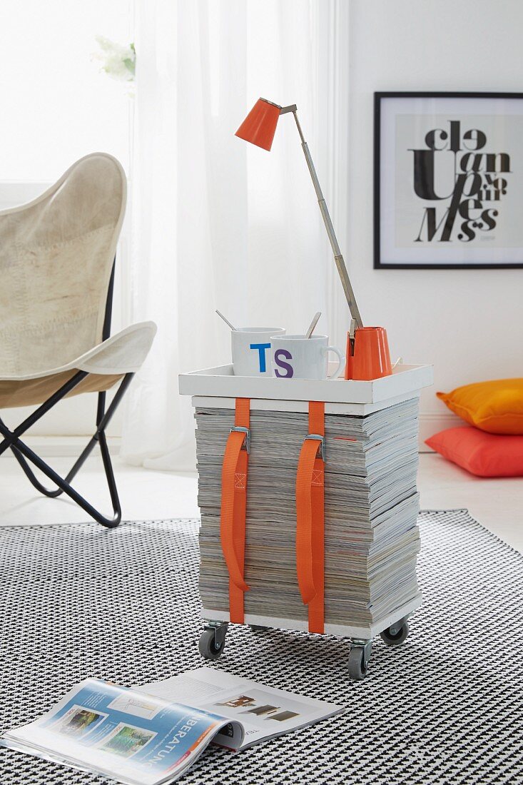 A table lamp and a cup of coffee on a mobile side table made from a stack of newspapers, wooden boards and straps