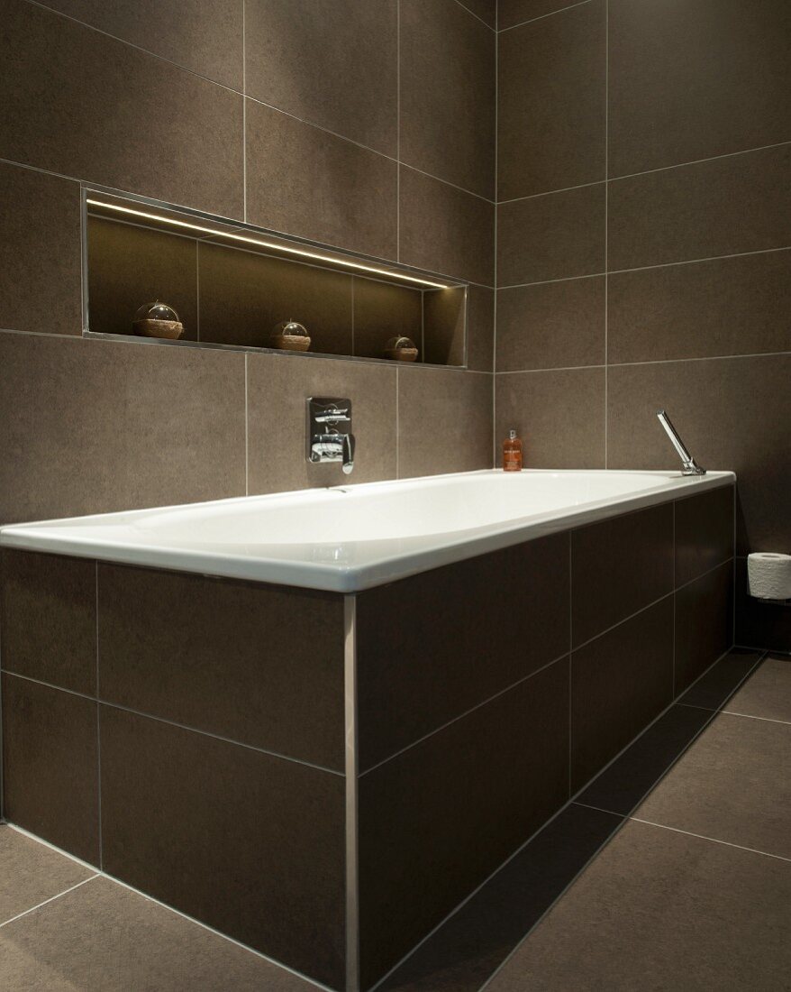 Braun gefliestes Badezimmer mit Badewanne und indirekt beleuchteter Ablagenische