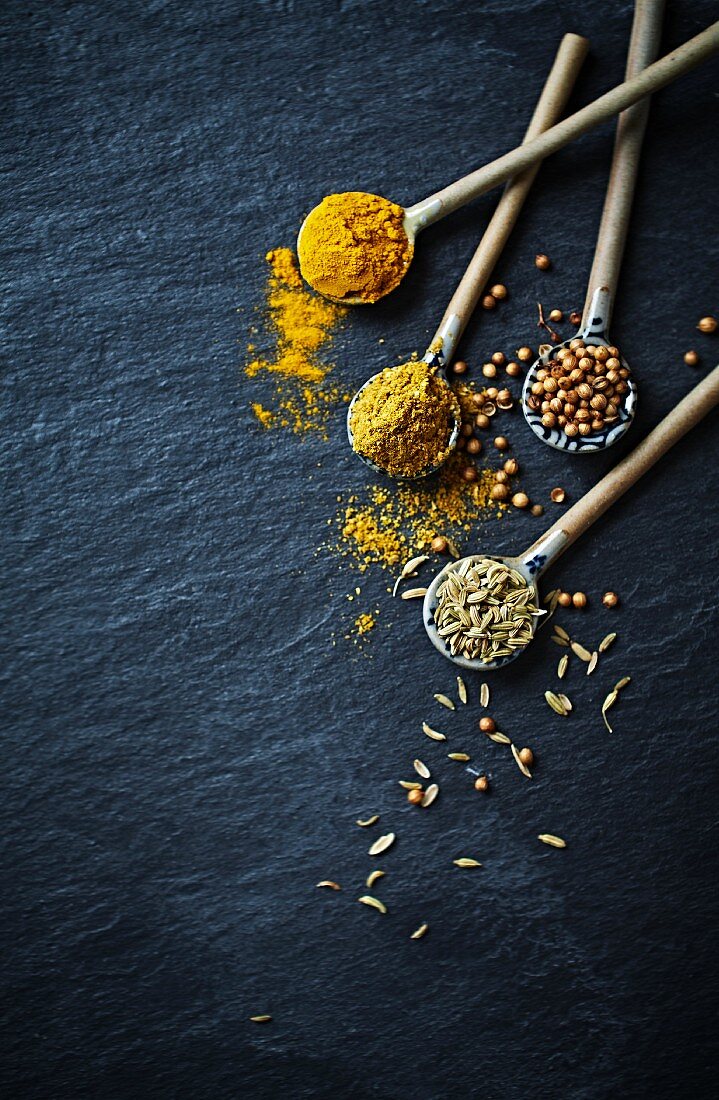 Curry powder, turmeric, cumin seeds and coriander seeds on ceramic spoons