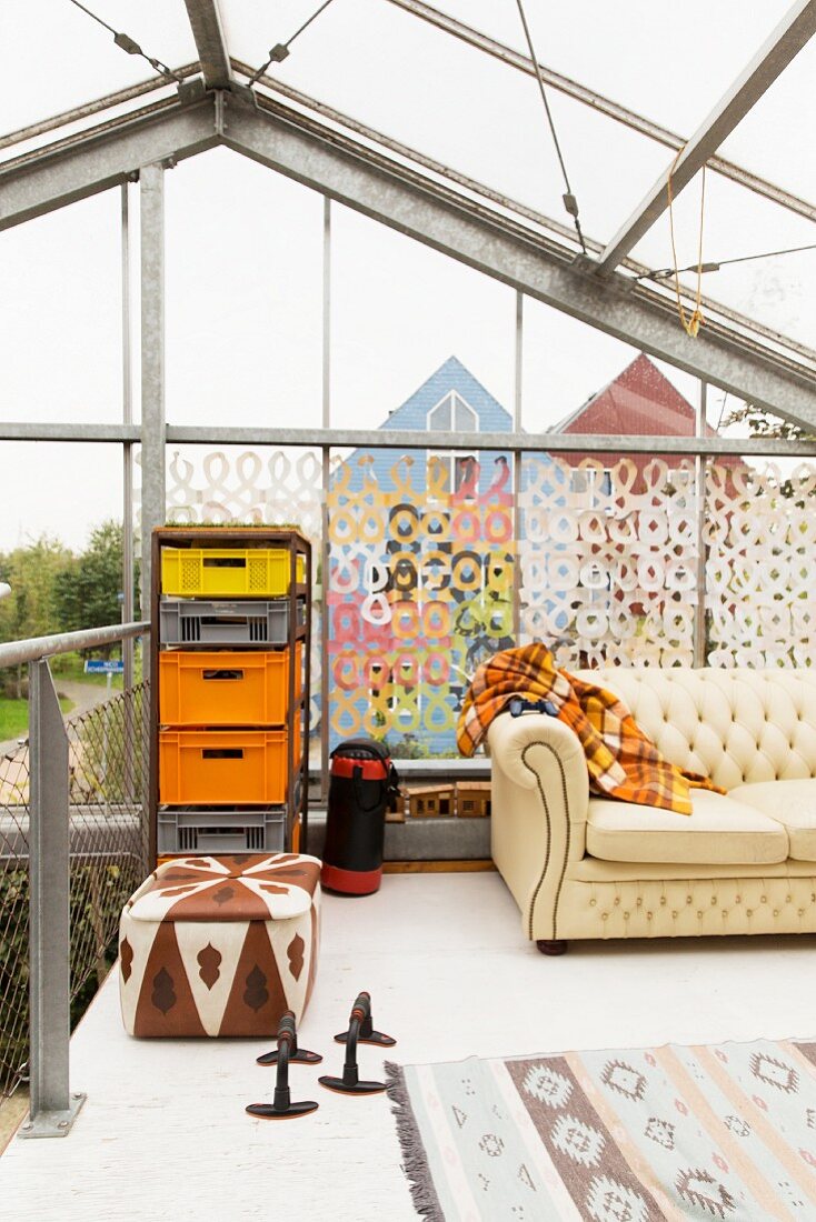 Patterned leather pouffe and metal frame holding colourful plastic crates against decorated glass façade on gallery in former greenhouse