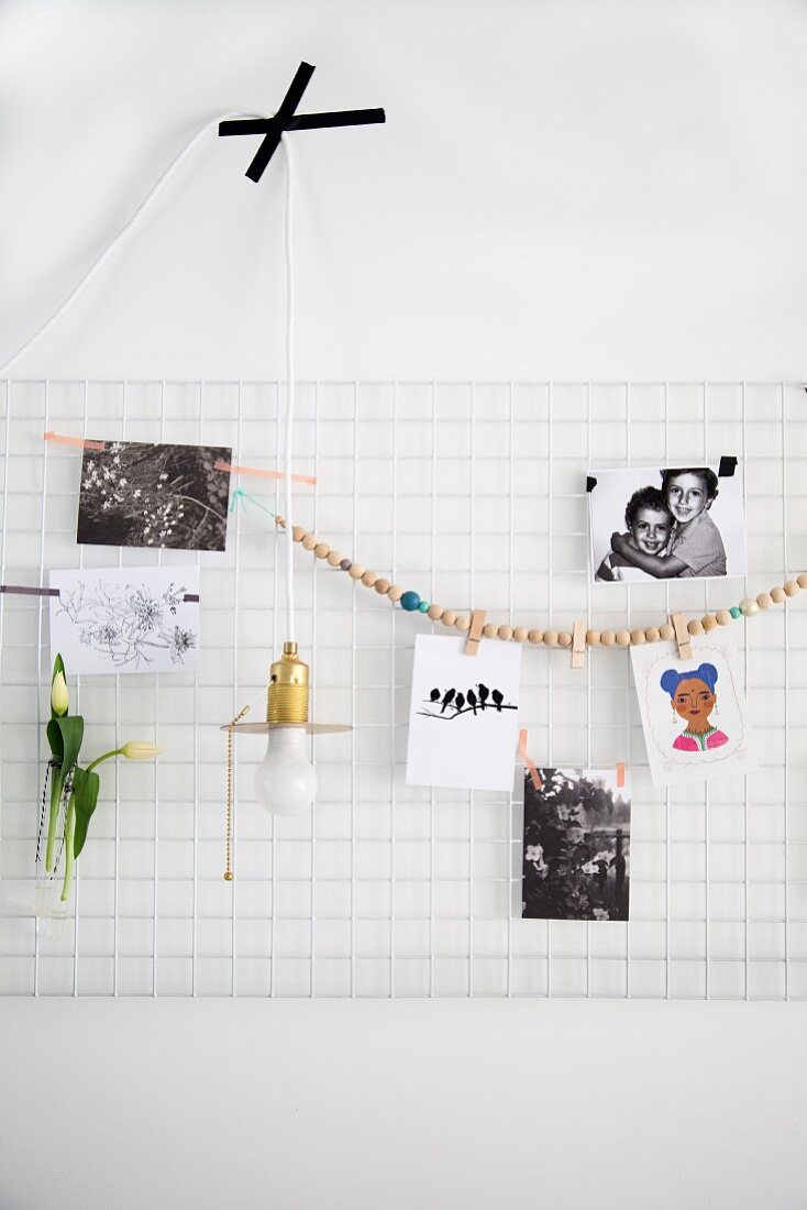 Photos and bead necklace hung on white metal mesh used as pinboard and minimalist pendant lamp stuck on wall with black tape