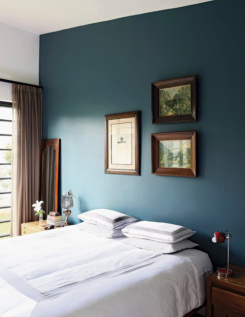 Bett mit weisser Bettwäsche vor petrolfarbener Wand und gerahmte Bilder im Schlafzimmer