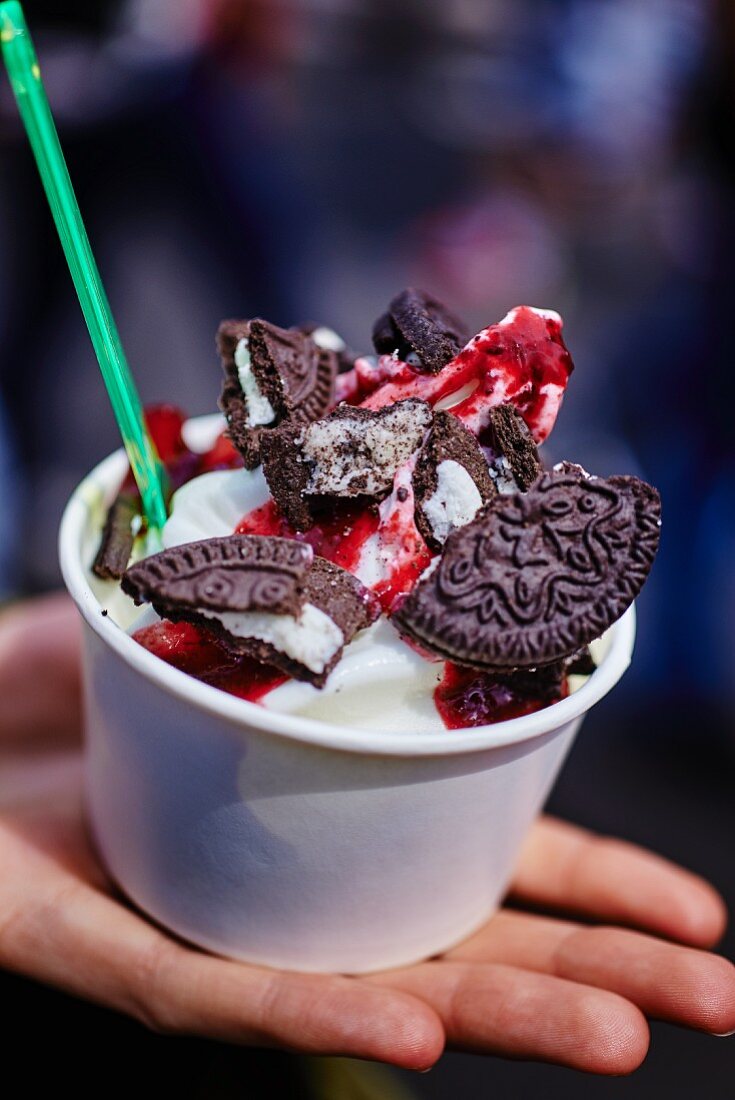 A hand holding a cup of frozen yoghurt with chocolate biscuits