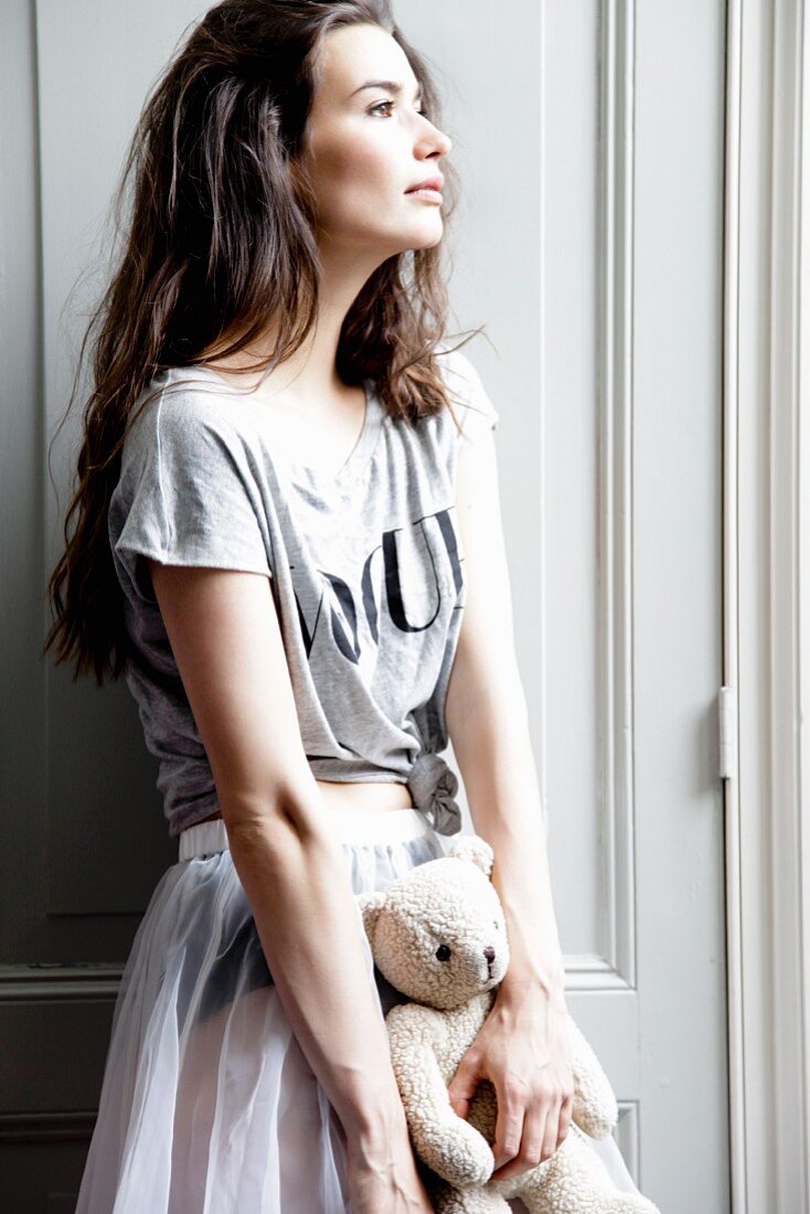 A young woman standing in front of a cupboard holding a teddy bear and wearing a T-shirt and a skirt