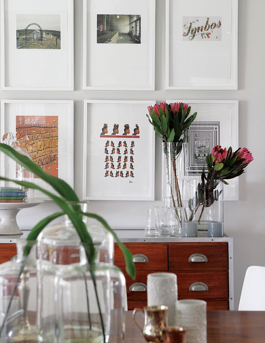 Leaves in vintage glass vessels, tropical flowers in glass vase on sideboard and gallery of pictures on wall