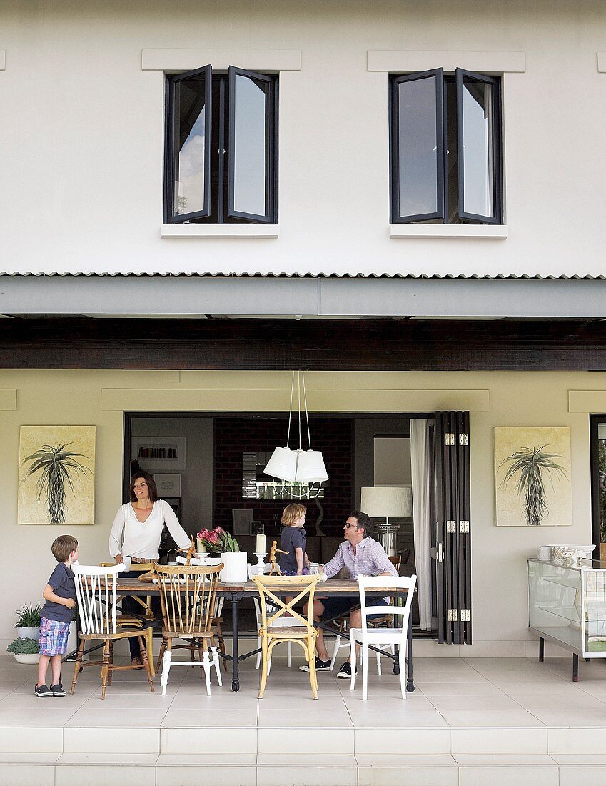 House with open, sliding, folding doors and roofed terrace; family sitting around table on various chairs