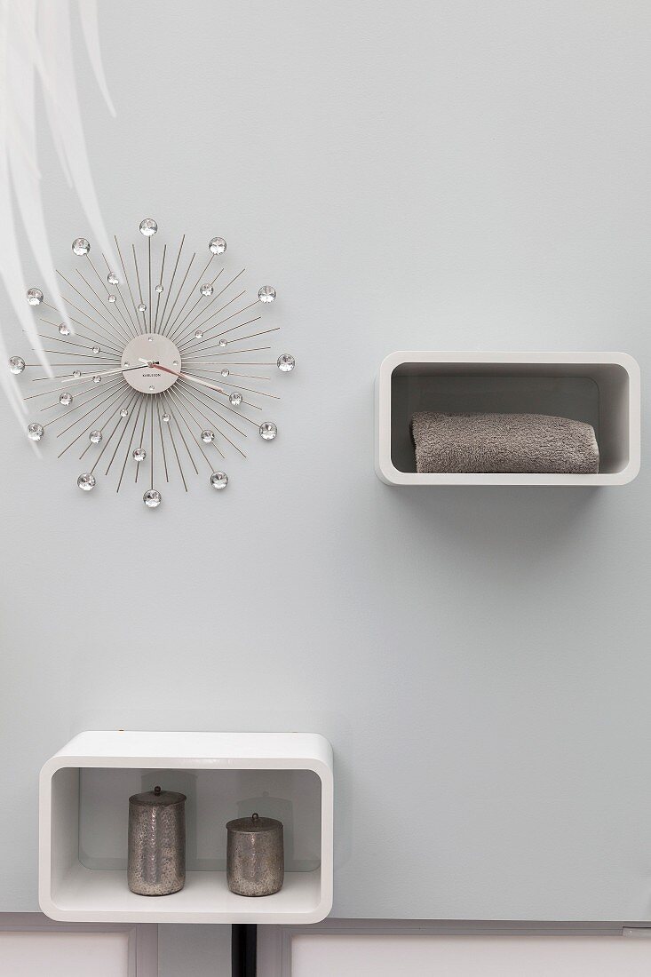 Elegant wall clock with crystal decorations and two white shelving units on pale wall