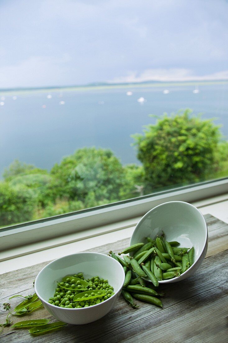 Peas and pea pods in bowls by a window with a sea view