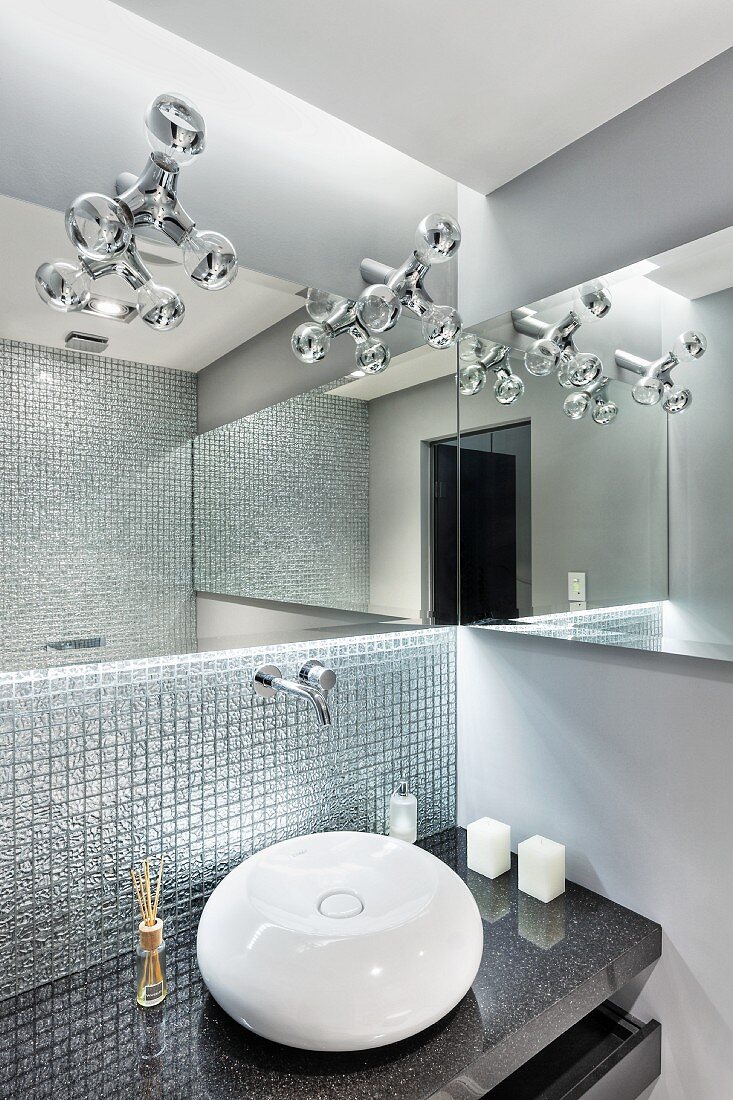 Spherical sink on granite counter against mosaic-tiled wall below designer lamps