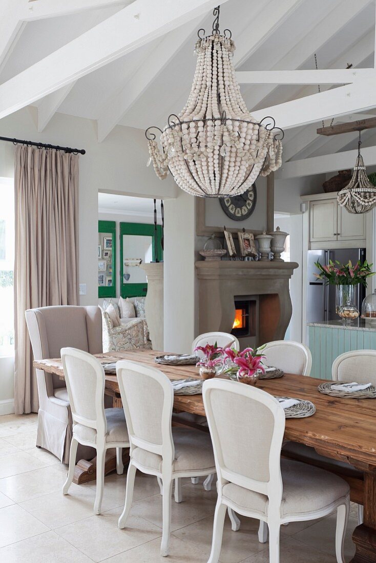 Rococo-style chairs around solid wooden table below antique chandelier and fireplace in background in open-plan interior with exposed roof structure