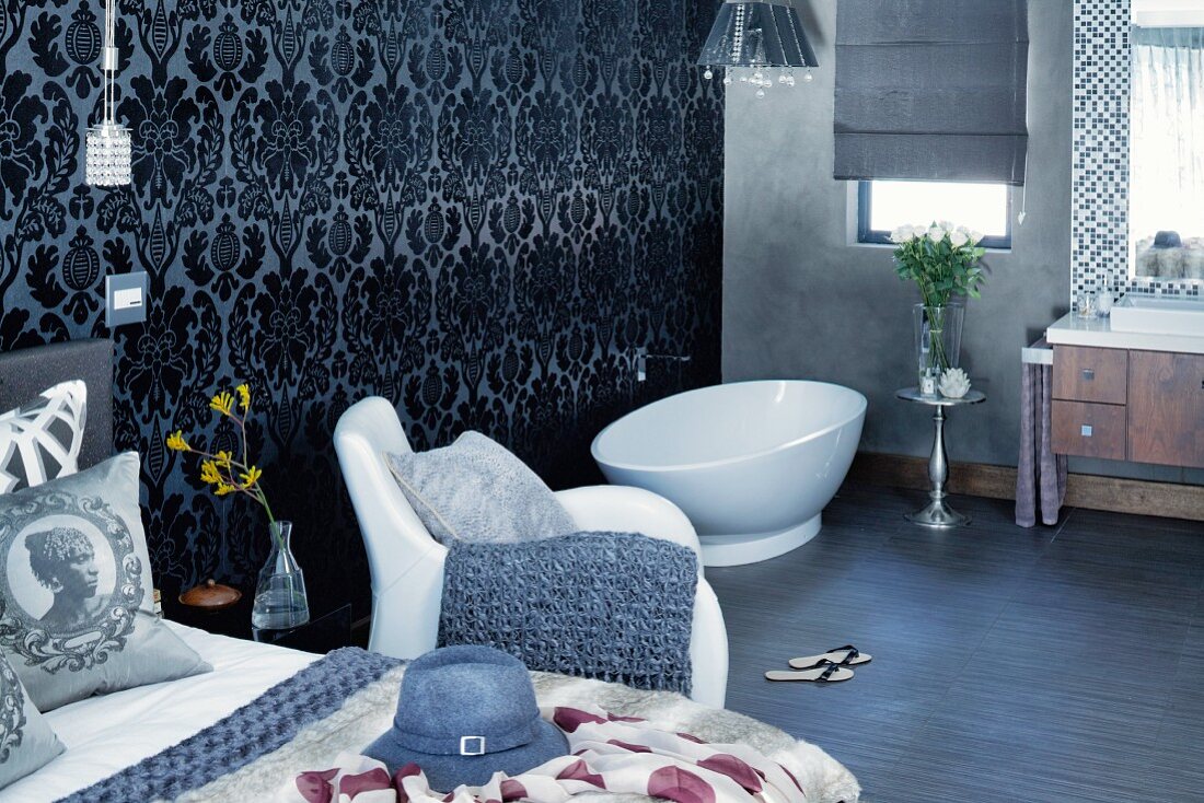 Bedroom with free-standing bathtub in open-plan ensuite bathroom, ornate wallpaper and white armchair next to bed