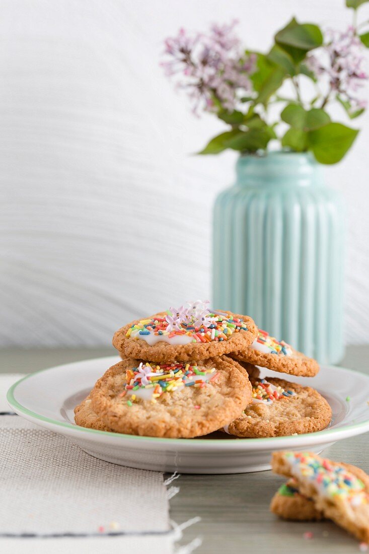 Oat biscuits with icing and sugar sprinkles with a vase of lilac flowers in the background