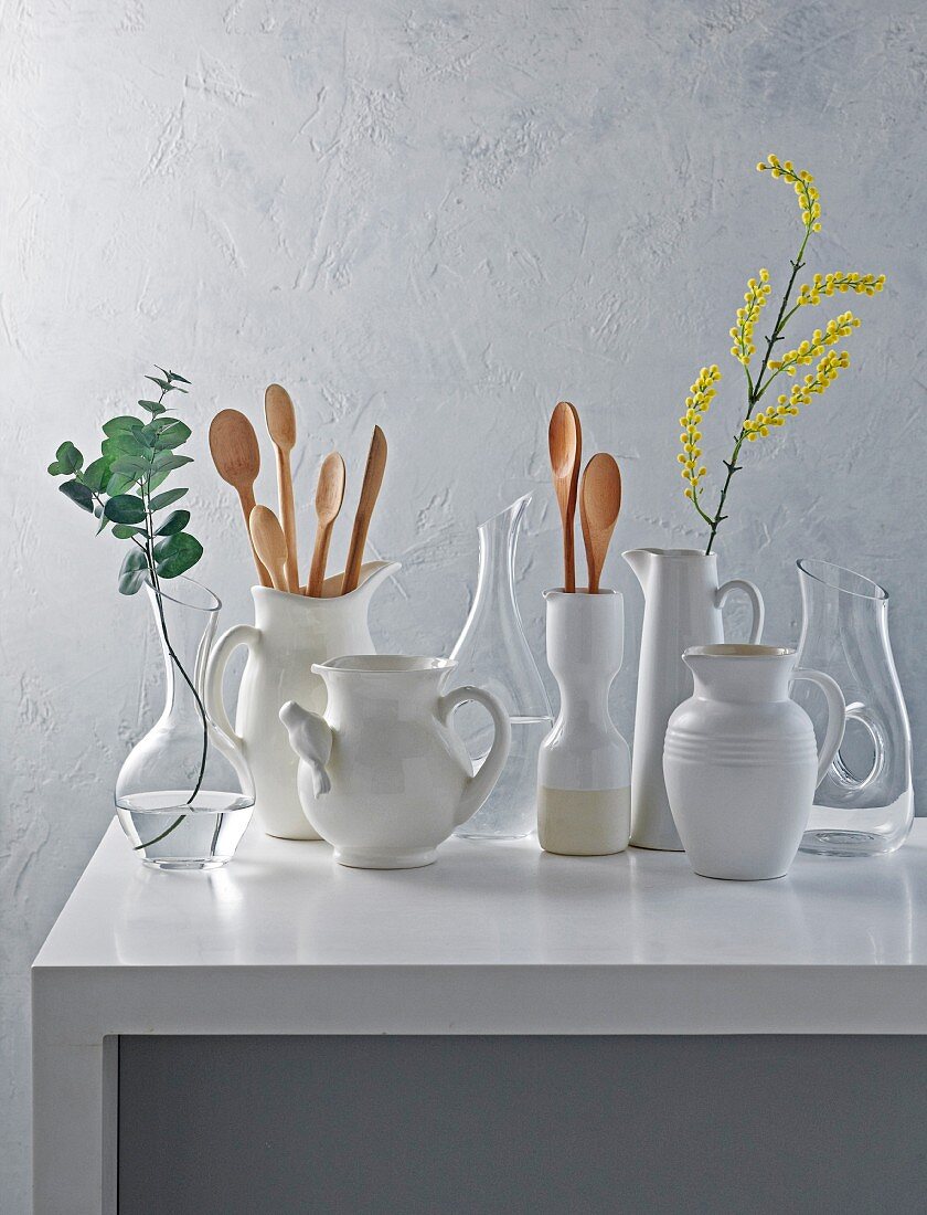 weiße Dekantierkannen und Keramikkrügen auf weißem Tisch, teilweise mit Blumenzweigen und Kochlöffeln bestückt