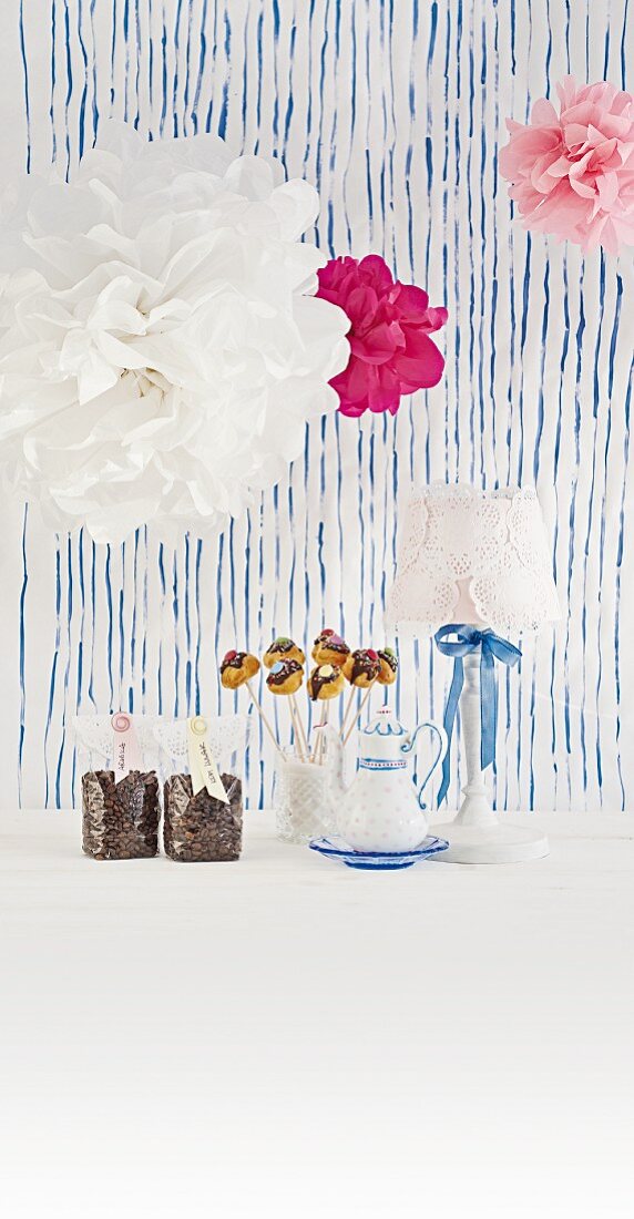 Ideen für den Kaffeetisch - Deko Pompons, Tischlampe mit Tortenspitzen, Cake Pops und verpackte Kaffeebohnen vor blauem Streifenmuster