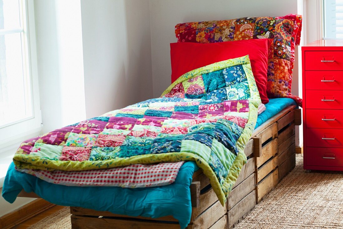 Rustic bed made from wooden crates with colourful patchwork blankets next to bright red metal filing cabinet